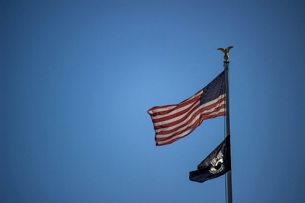 Photograph of American flag and POW flag at half mast. Blue sky in background